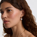 A close-up of a woman with wavy brown hair wearing a delicate gold hoop earring with a clear round gemstone drop, shown against a plain light background.