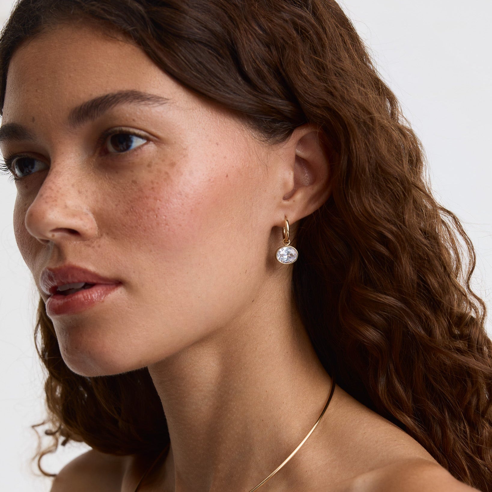 A close-up of a woman with wavy brown hair wearing a delicate gold hoop earring with a clear round gemstone drop, shown against a plain light background.