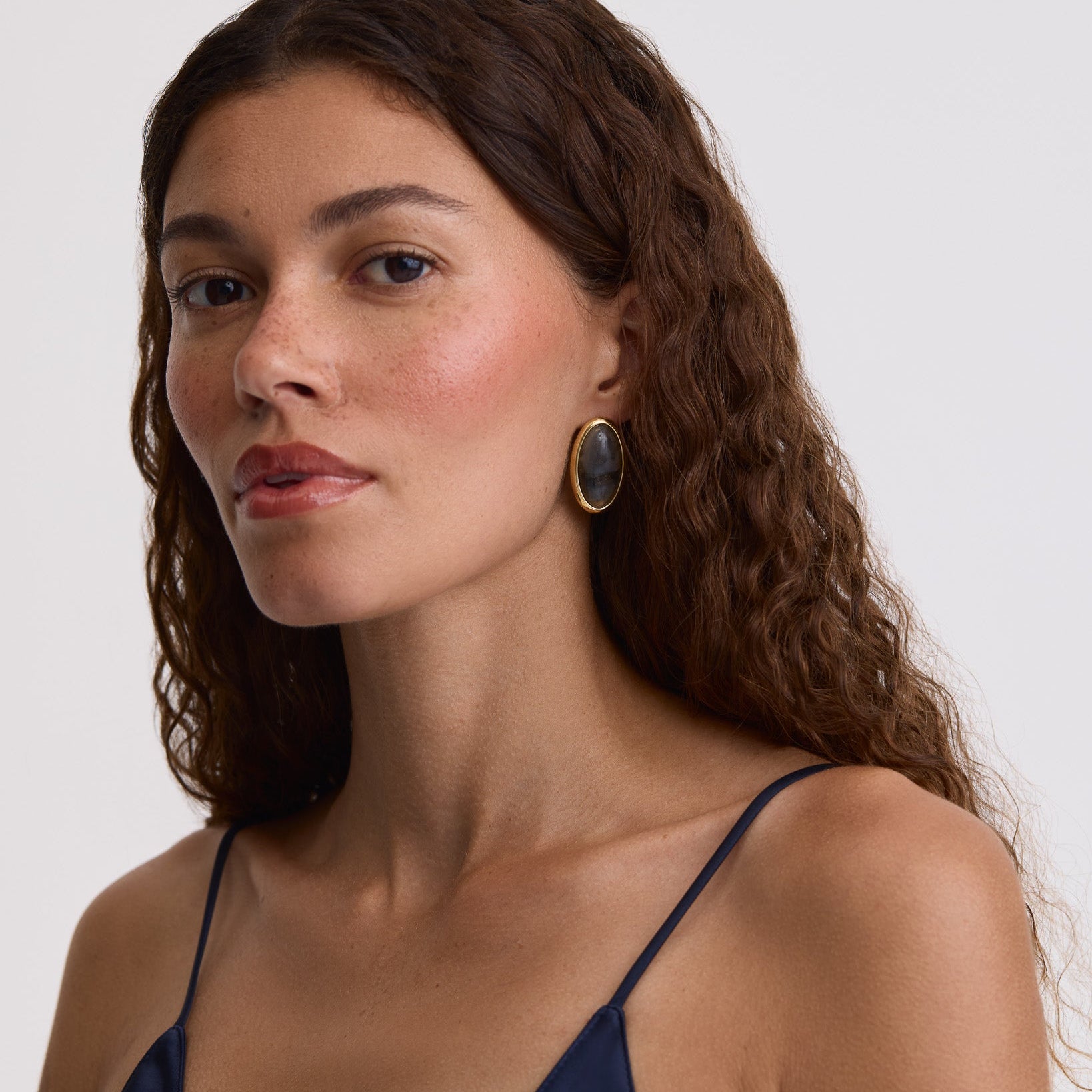 A close-up portrait of a woman with long, wavy brown hair wearing a dark navy blue spaghetti-strap top and a single oval gold earring with a dark stone, set against a plain light gray background.