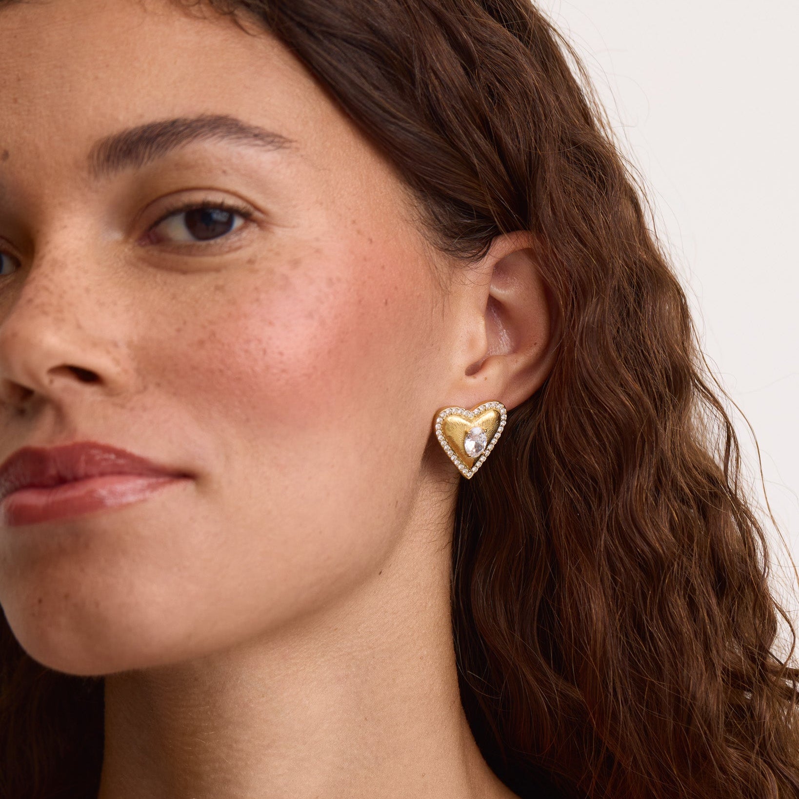 A close-up of a woman wearing a gold heart-shaped stud earring with a white textured center and small clear stones around the edge, against a plain light background.