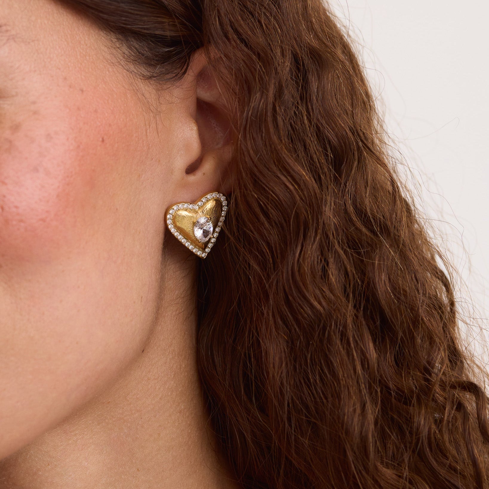 A close-up of a model's ear wearing a gold heart-shaped stud earring with a clear central gemstone and a border of small clear stones, set against a light neutral background.