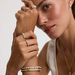 A close-up lifestyle shot of a model wearing multiple gold jewelry pieces, including a ring with a large clear gemstone, several delicate rings, a bracelet, and a necklace, against a clean light gray background.