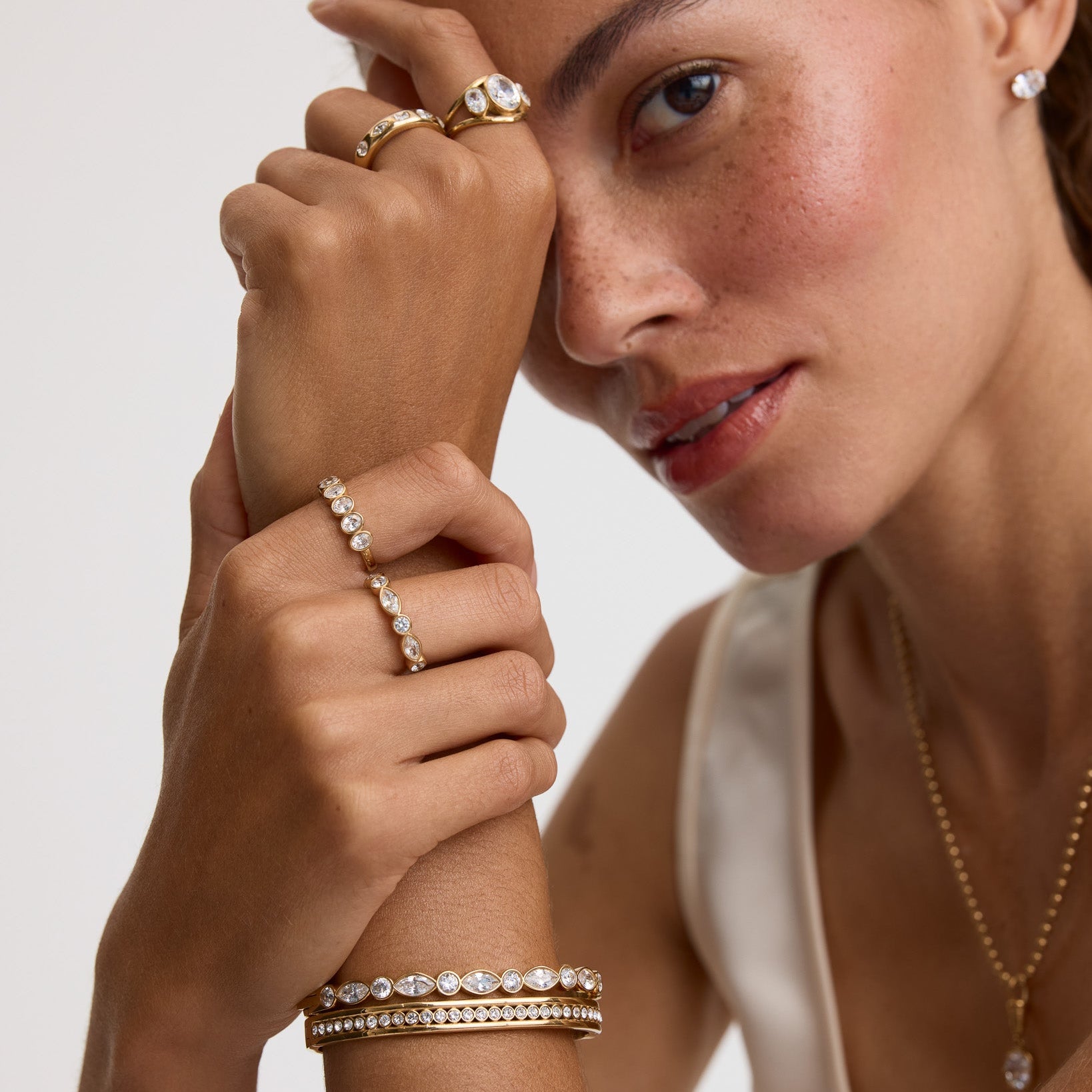 A close-up lifestyle shot of a model wearing multiple gold jewelry pieces, including a ring with a large clear gemstone, several delicate rings, a bracelet, and a necklace, against a clean light gray background.