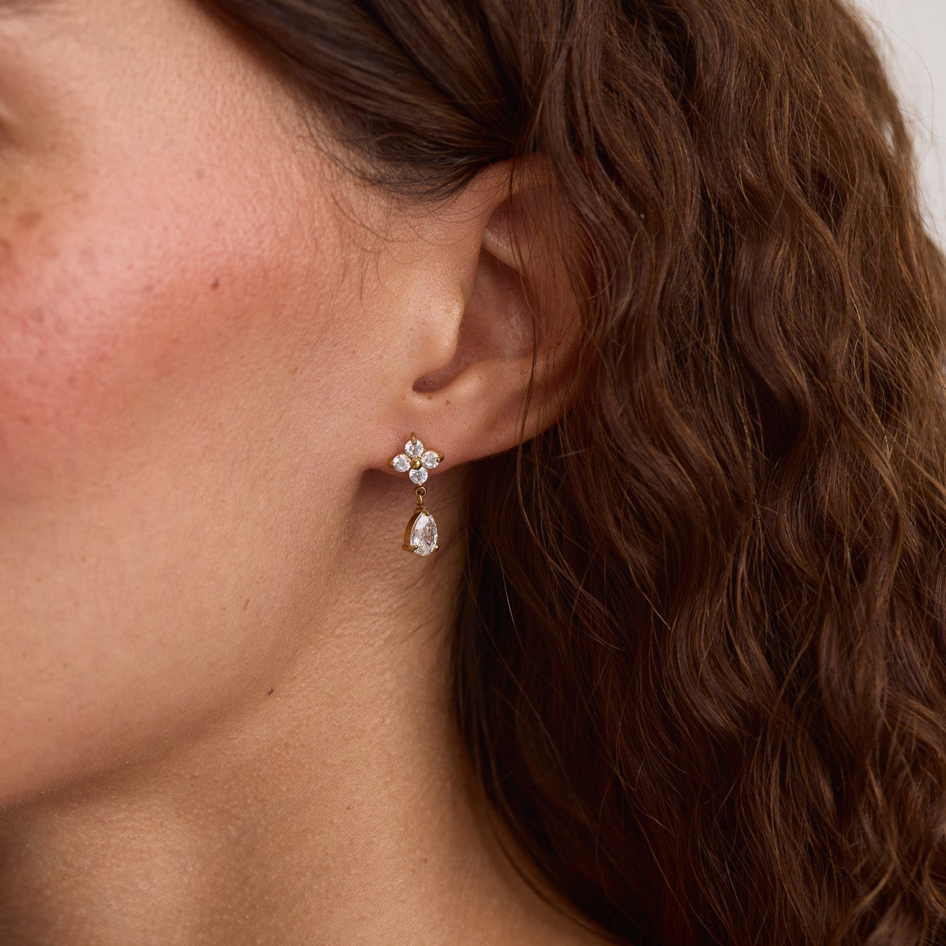 A close-up of a woman's ear wearing a small floral stud earring with a dangling clear teardrop stone, shown against her hair and skin.