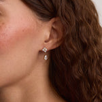 A close-up of a woman's ear wearing a small floral stud earring with a dangling clear teardrop stone, shown against her hair and skin.