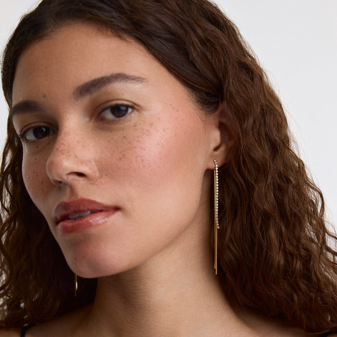 A close-up of a woman with wavy brown hair wearing a long, delicate gold dangle earring with a small clear stone cluster at the top.