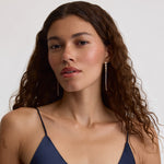 A woman with long, wavy brown hair wearing a navy blue satin camisole top and long, dangling gold-toned earrings, photographed against a clean, light gray studio background.