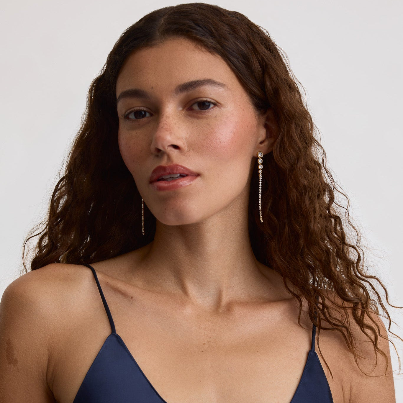 A woman with long, wavy brown hair wearing a navy blue satin camisole top and long, dangling gold-toned earrings, photographed against a clean, light gray studio background.