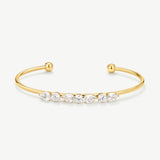 A gold open cuff bracelet featuring a row of clear marquise and round-cut gemstones across the front, displayed on a clean off-white background.