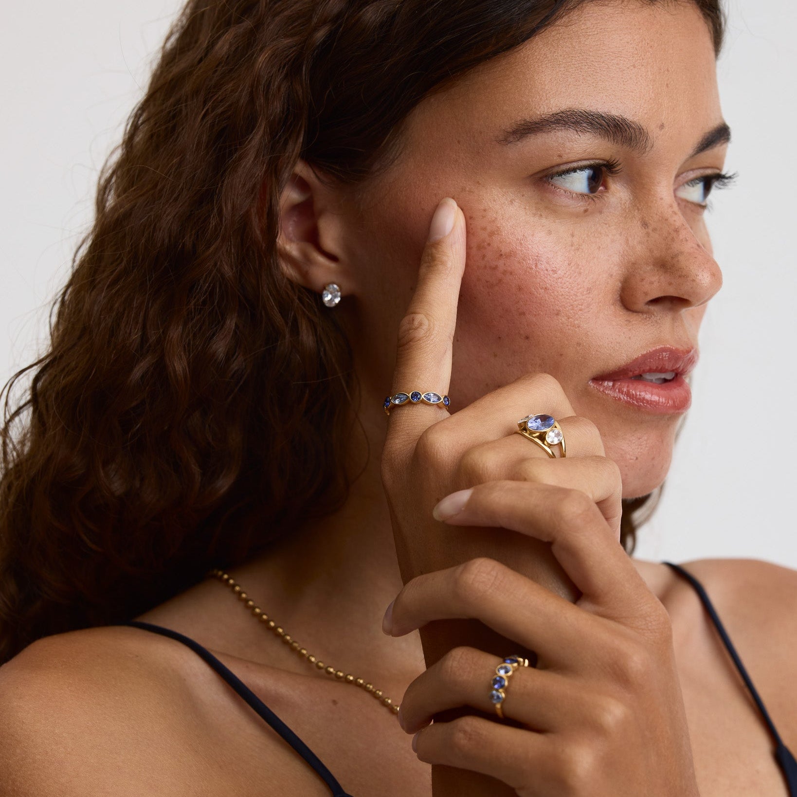 A close-up lifestyle shot of a woman with curly brown hair wearing multiple gold rings with blue gemstones and a gold chain necklace, touching her cheek with her index finger against a light gray background.