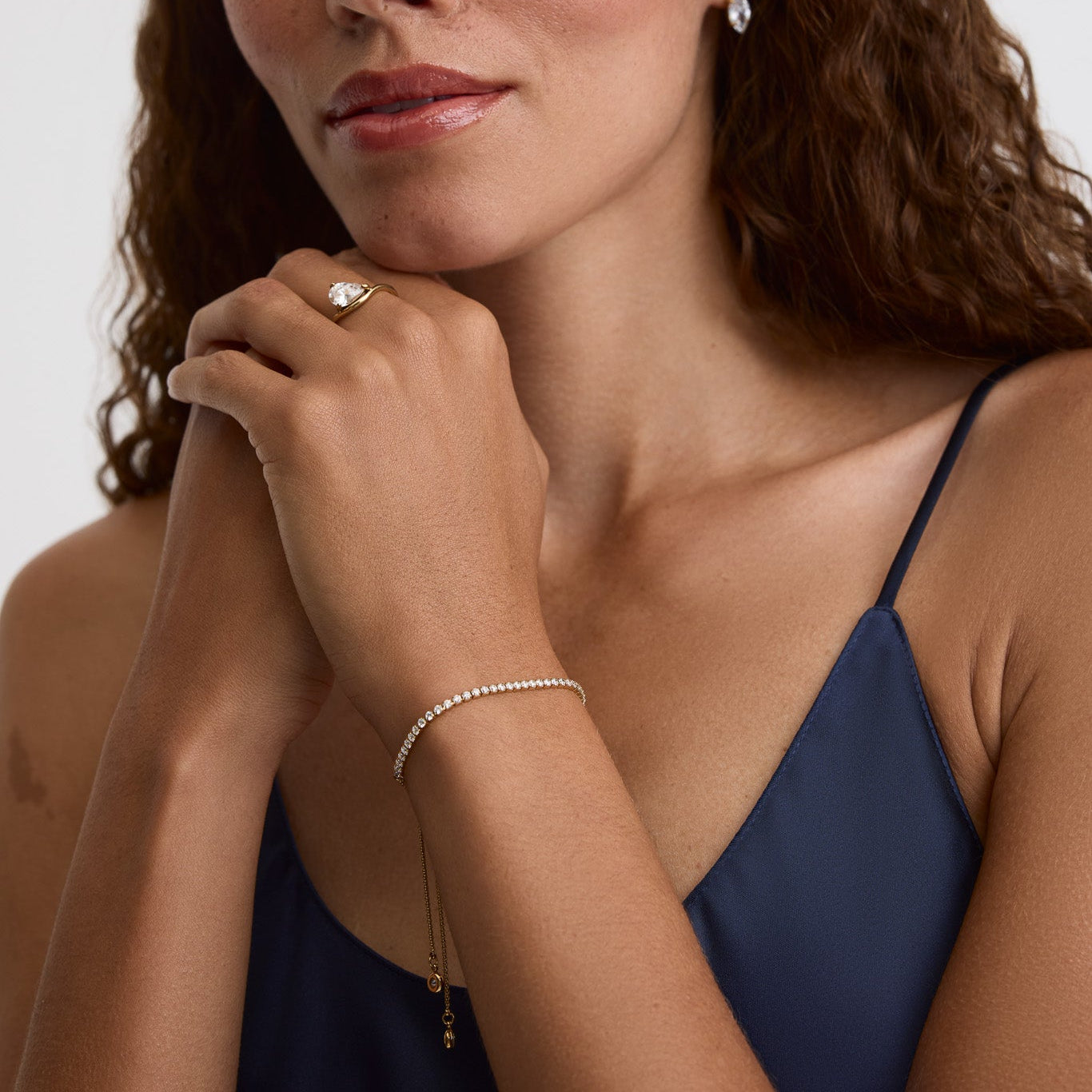 A close-up portrait of a woman with long brown hair wearing delicate jewelry, including a small silver hoop earring, a thin silver bracelet, and a ring with a clear gemstone, against a plain light gray background.