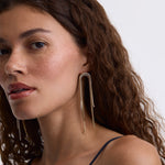 A close-up of a woman with curly brown hair wearing long, dangling gold-toned earrings with a textured, beaded look against a plain light background.