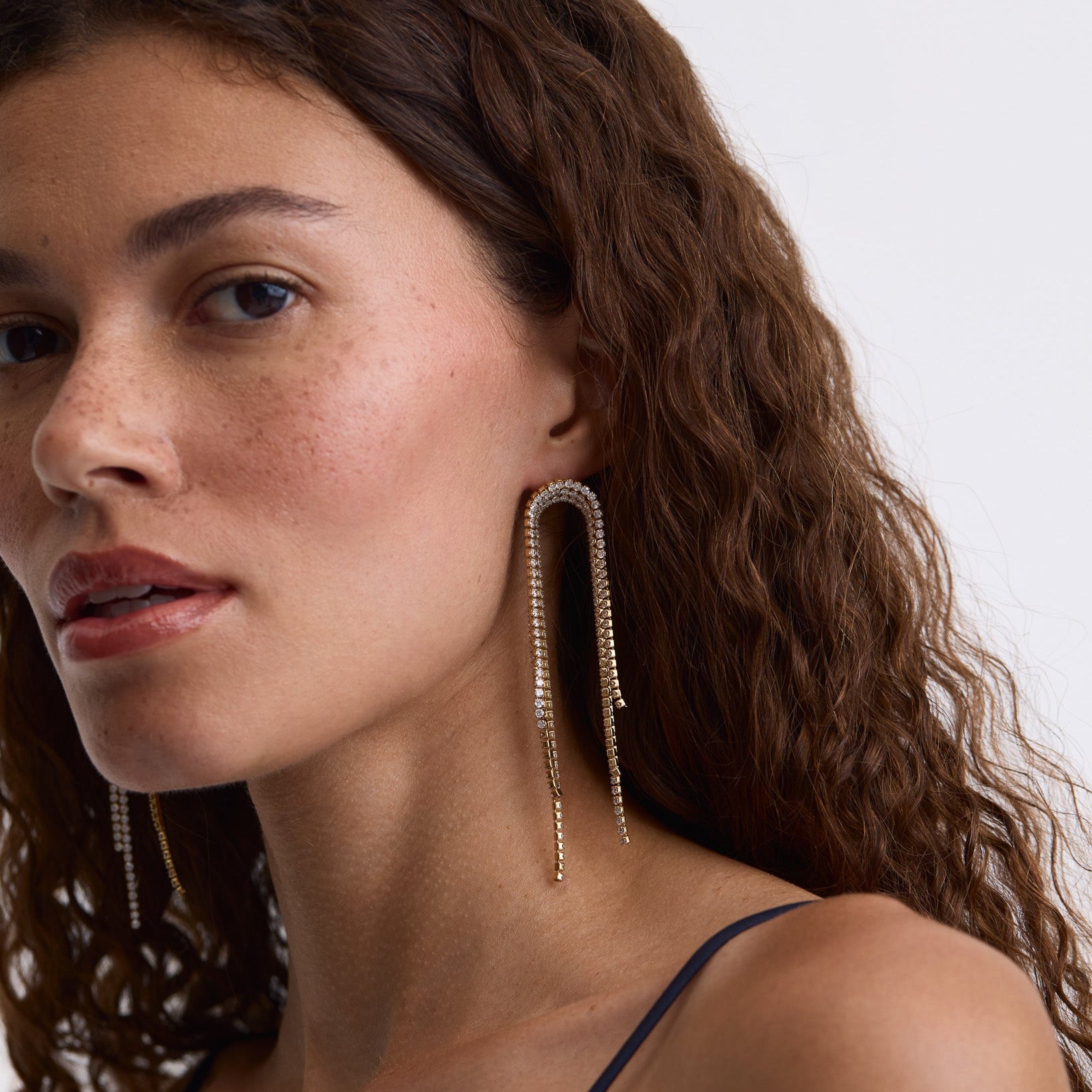 A close-up of a woman with curly brown hair wearing long, dangling gold-toned earrings with a textured, beaded look against a plain light background.