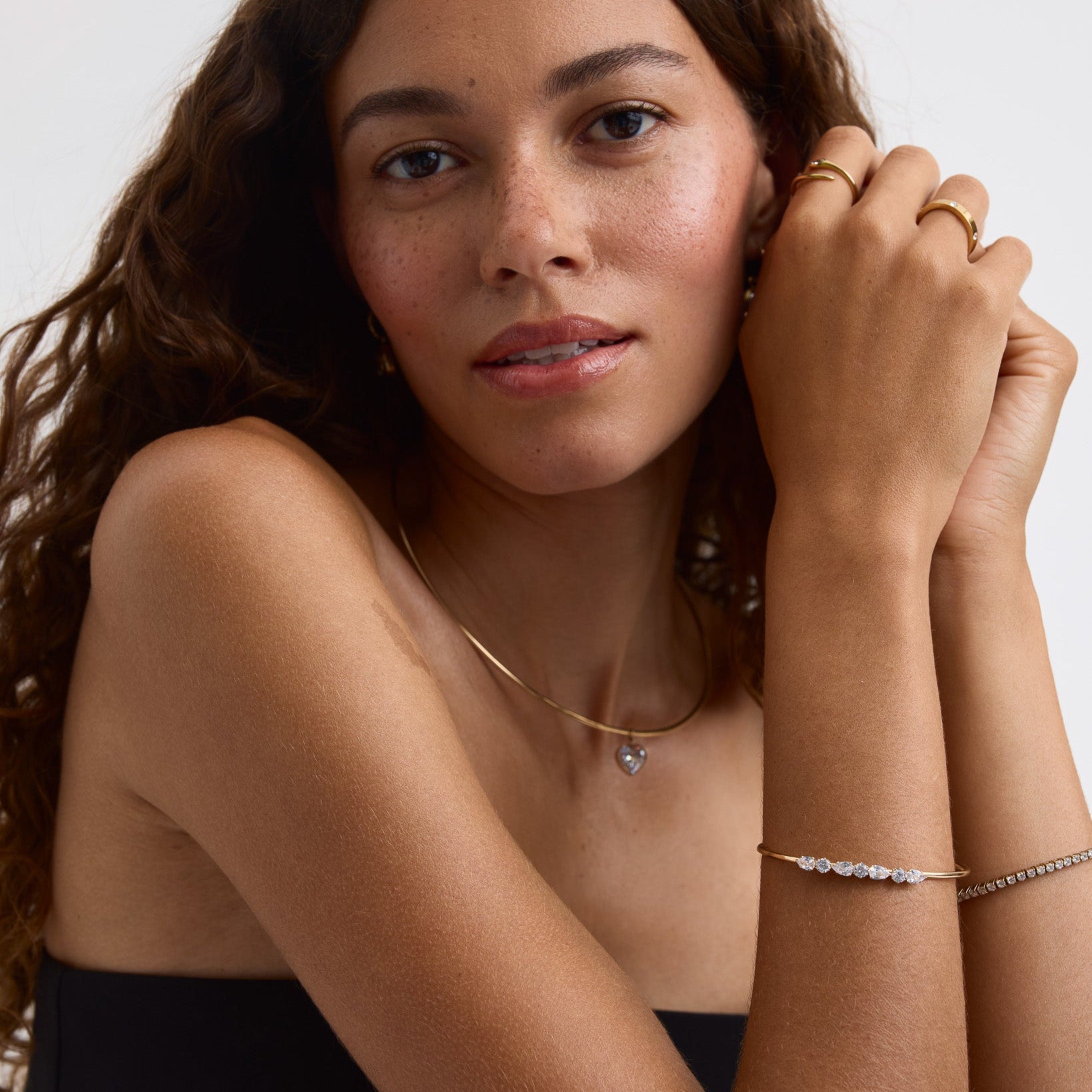 A close-up of a woman with long wavy brown hair wearing delicate gold jewelry, including a thin gold chain necklace with a small heart pendant and gold rings on her fingers, against a plain light background.