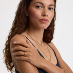 A woman with long, wavy brown hair is shown from the chest up against a plain light background, wearing a delicate gold chain necklace, a gold ring with a clear gemstone, and a gold bracelet.