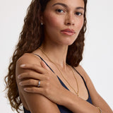 A woman with long, wavy brown hair is shown from the chest up against a plain light background, wearing a delicate gold chain necklace, a gold ring with a clear gemstone, and a gold bracelet.