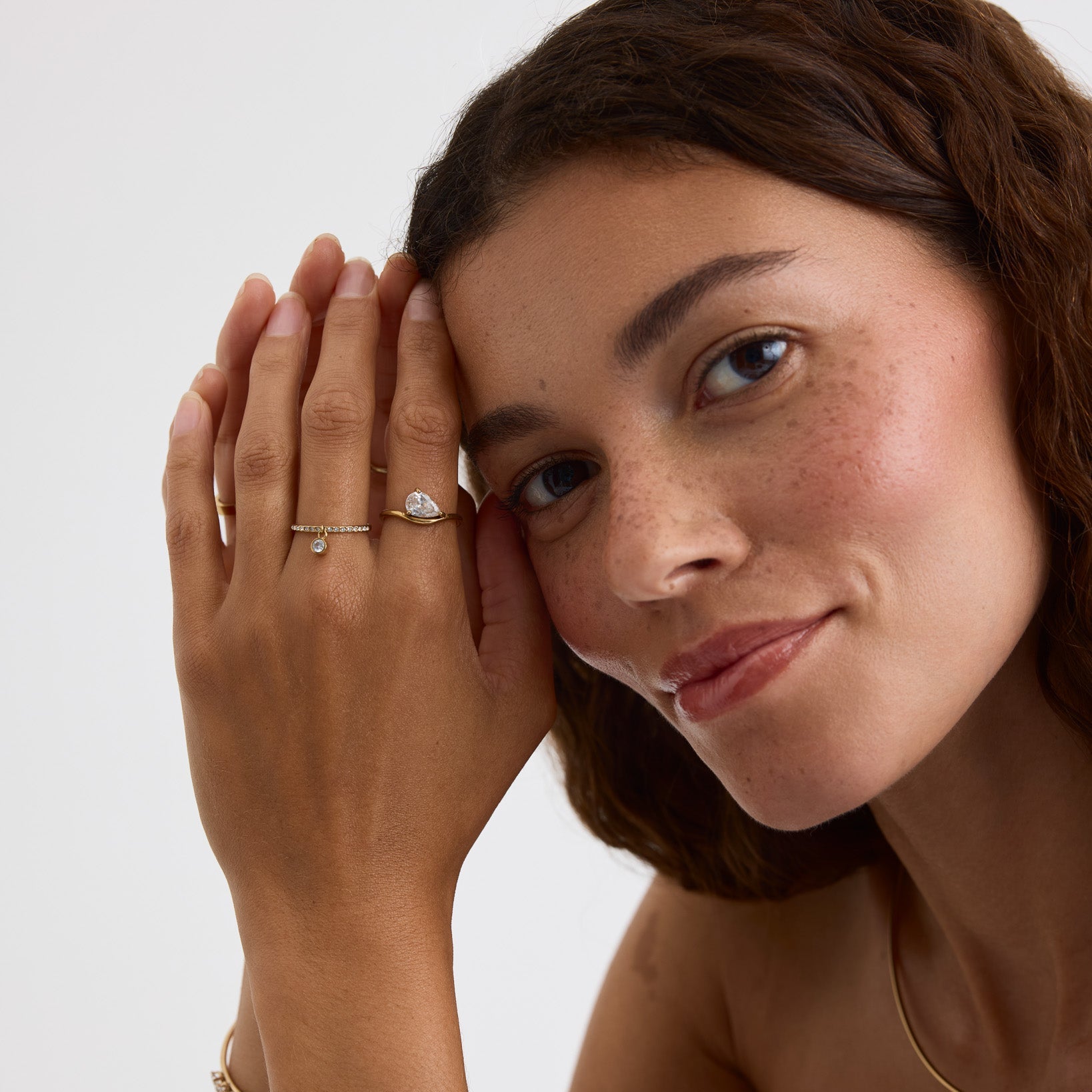 A close-up lifestyle shot of a woman with her hands resting against her face, showcasing delicate jewelry including a silver ring with a clear gemstone, a silver band with tiny clear stones, a gold bracelet, and a gold necklace with a heart charm, set against a clean light gray background.