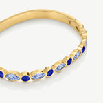 A close-up of a gold bangle bracelet featuring alternating round and marquise-cut blue gemstones in gold bezel settings, shown on a clean white background.