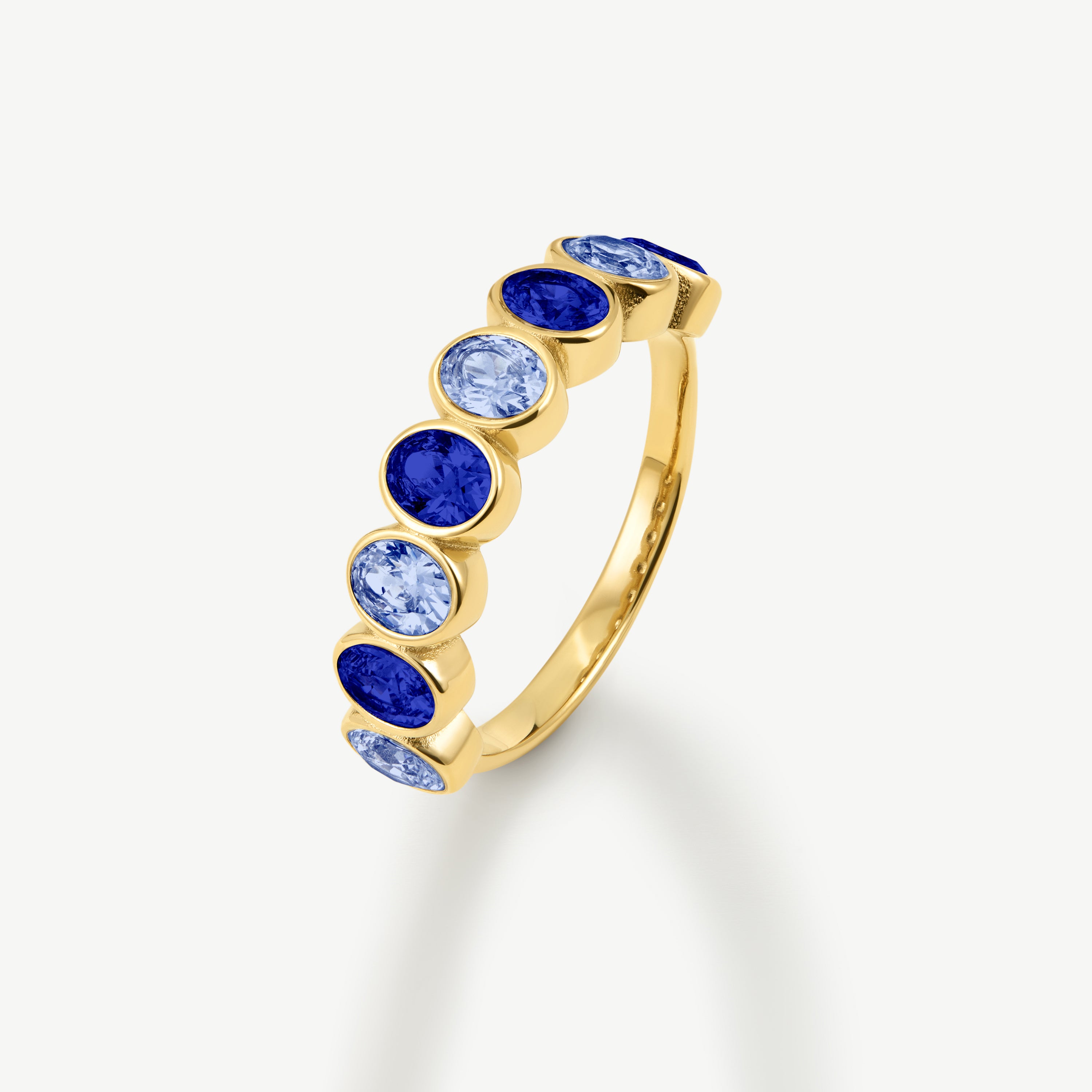 A gold band ring featuring a row of oval-cut blue and light blue gemstones, shown on a clean off-white background.