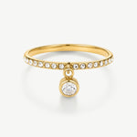 A gold ring featuring a thin band paved with small clear round stones and a dangling bezel-set round clear gemstone, shown on a clean white background.