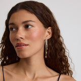 A close-up of a woman with curly brown hair wearing long, dangling silver-toned earrings with small clear stones, paired with a navy camisole top against a light gray background.