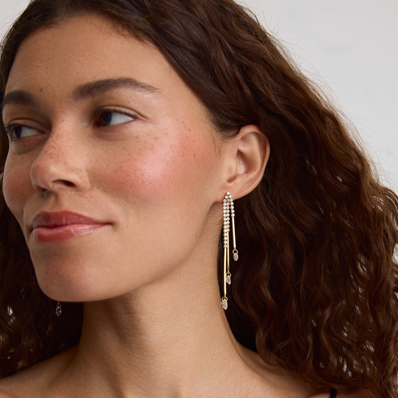 A close-up of a woman wearing a long, dangling gold-toned beaded earring, shown from the chest up against a light neutral background.