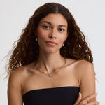 A woman with long, wavy brown hair is shown from the chest up against a light gray studio background. She is wearing a black strapless top and delicate gold jewelry, including a necklace with a small blue stone, stud earrings, and rings.