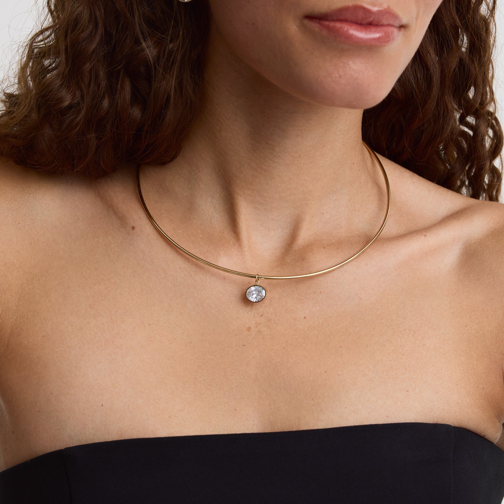 A close-up of a model wearing a delicate gold choker necklace with a small clear gemstone pendant, paired with a black strapless top.