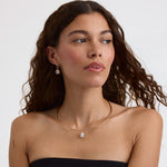 A woman with long, wavy brown hair is shown from the chest up against a light gray studio background. She is wearing a delicate gold necklace with a small teardrop-shaped clear gemstone pendant and matching drop earrings.