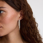 A close-up of a woman with wavy brown hair wearing a long, dangling silver earring with multiple rows of small clear stones against a plain light background.
