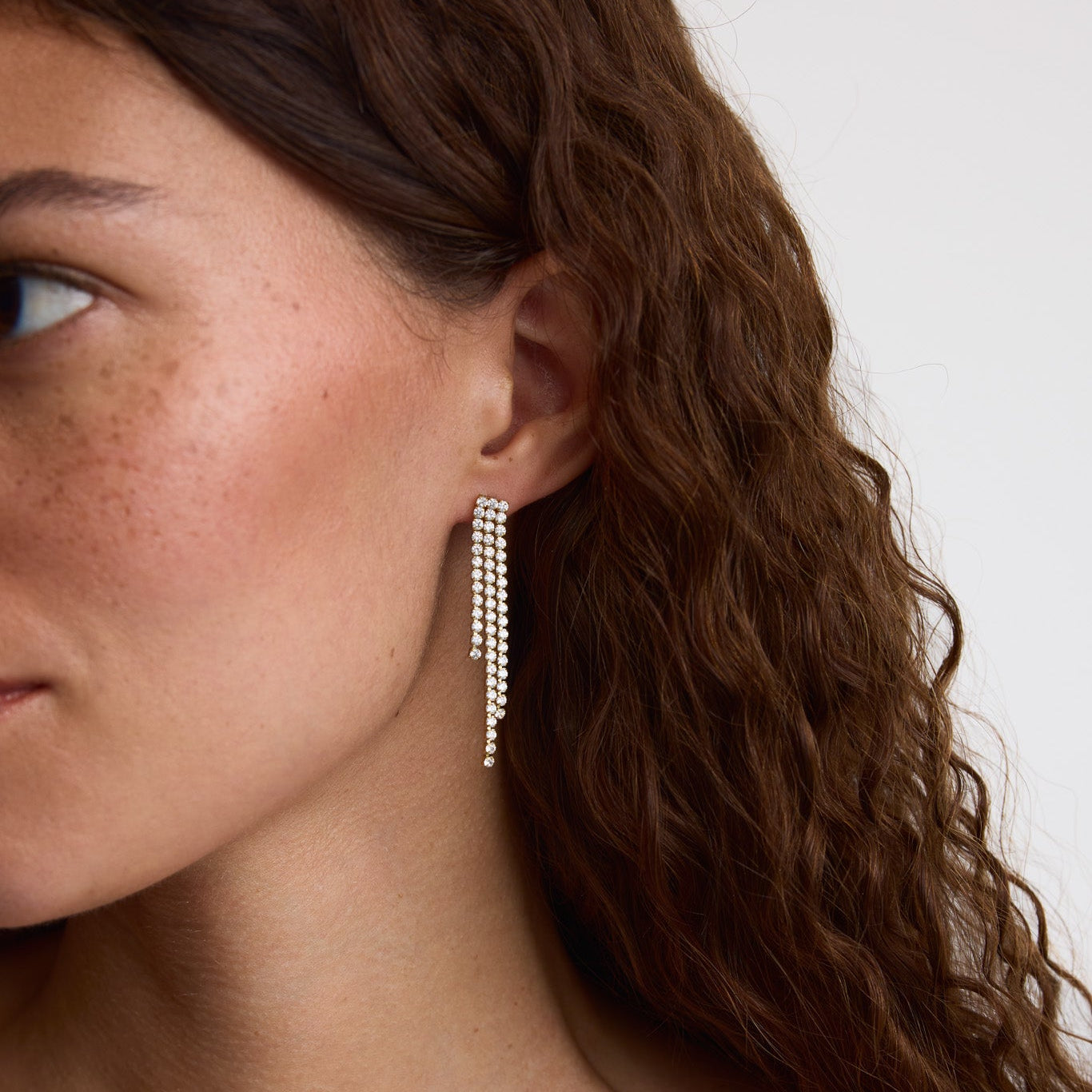 A close-up of a woman with wavy brown hair wearing a long, dangling silver earring with multiple rows of small clear stones against a plain light background.