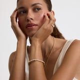 A close-up portrait of a woman with her hands resting on her cheeks, wearing delicate gold jewelry including a gold chain necklace, a gold bracelet, and a gold ring with a clear gemstone, set against a clean white background.
