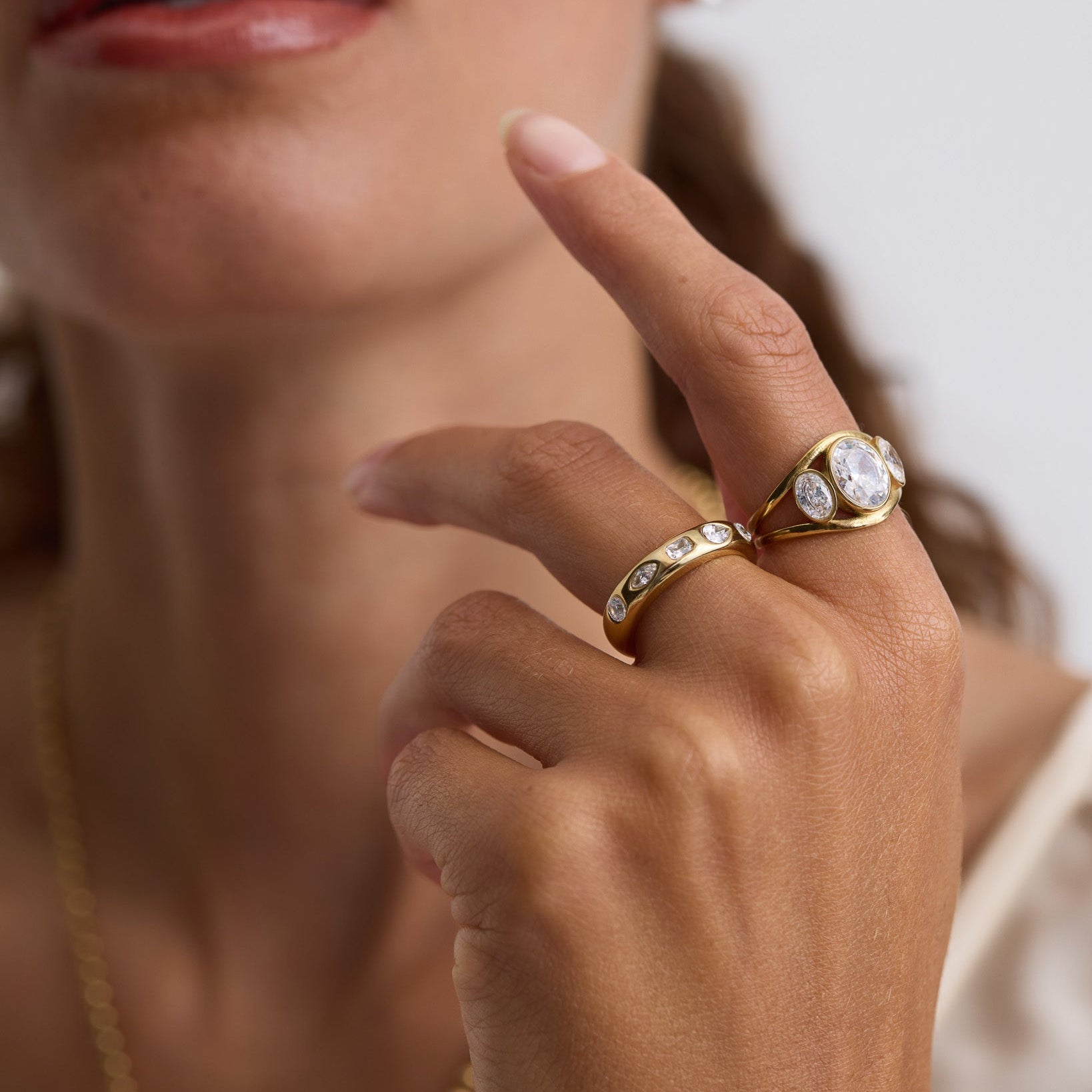 A close-up lifestyle shot of a model wearing two gold rings. The primary ring features a large oval-cut clear gemstone in a bezel setting, while the band ring has a channel-set pattern of small clear round gemstones.